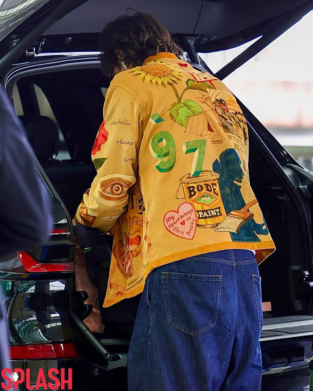 Мужчина с яркой курткой и джинсами наклоняется к багажнику автомобиля, из ушей торчат белые проводные наушники — кадр фиксирует повседневный момент