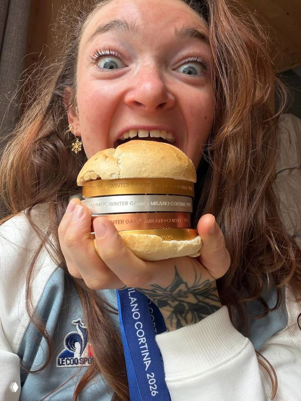 Французская биатлонистка Лу Жанмонно улыбается и держит «бургер» из булочки и стопки медалей Milano Cortina 2026, позируя для снимка.