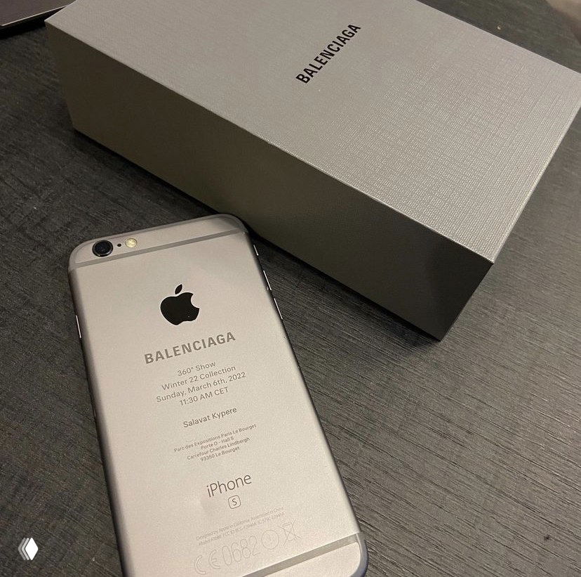 Серебристый iPhone с выгравированными деталями показа Balenciaga рядом с коробкой бренда на тёмной поверхности — креативное приглашение в виде телефона.