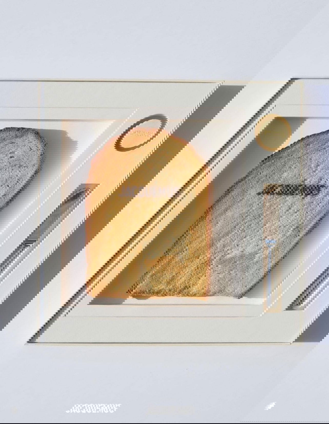 Ломтик тоста с отпечатанным логотипом Jacquemus, поданный в коробке с маслом и ножом на светлом фоне — концептуальное приглашение‑завтрак.