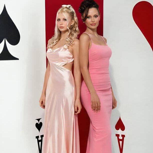 Полноразмерный кадр: две женщины в вечерних платьях позируют на фоне декоративных карт, элегантные позы и розовые оттенки в образах.