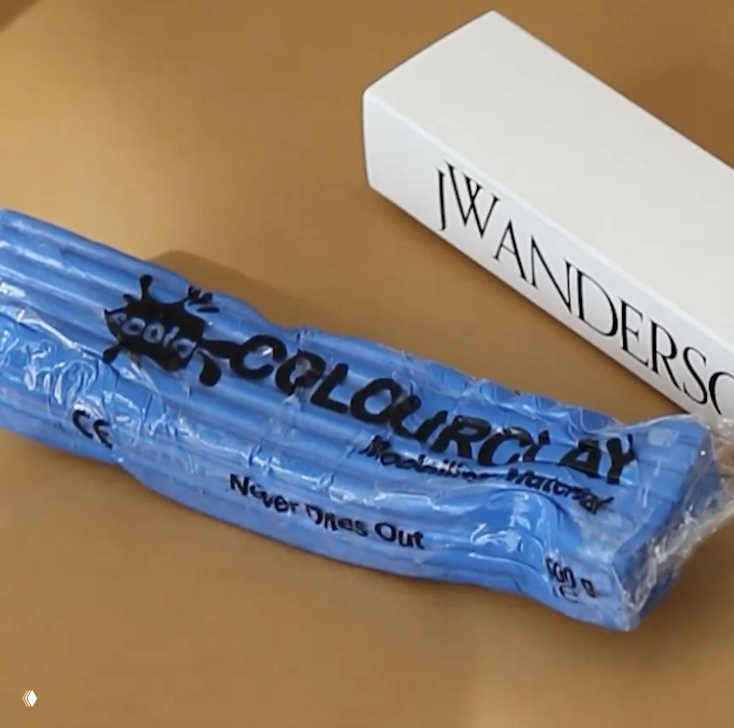 Пачка синего пластилина в прозрачной упаковке лежит рядом с коробкой JW Anderson — небольшая скульптурная реквизитная идея для приглашения на показ.