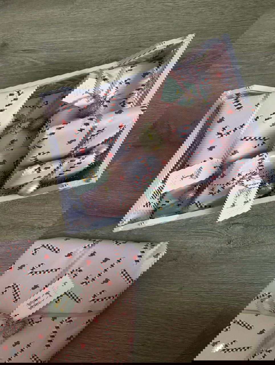 Ящик с множеством розовых подарков, завернутых в бумагу с узором тростей и сердечек, украшенных бирками и небольшими ёлочными шариками.