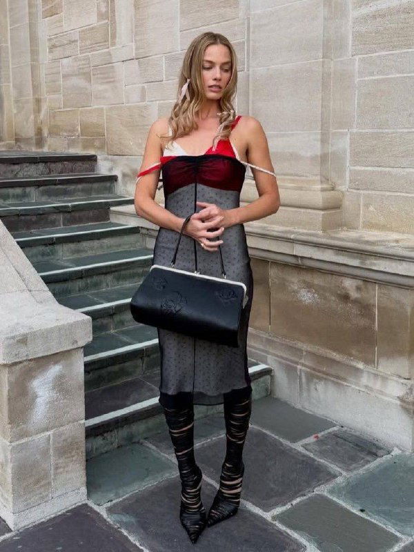 Прозрачное чёрно‑красное платье‑слип с тонкими бретелями и крупной чёрной сумкой на ступенях у каменной стены в дневной съёмке.
