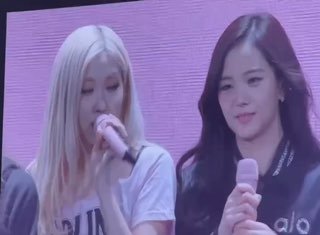 Крупный план двух участниц BLACKPINK на сцене во время финала тура, они поют в микрофоны на фоне розового экрана, видно эмоциональное выражение