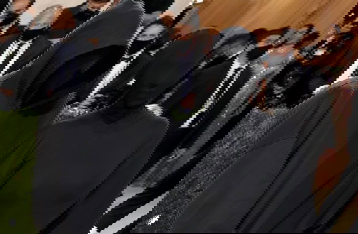 Два человека в чёрных плотных образах с закрытыми лицами и капюшонами на красной дорожке, вокруг — фотографы и гости мероприятия.