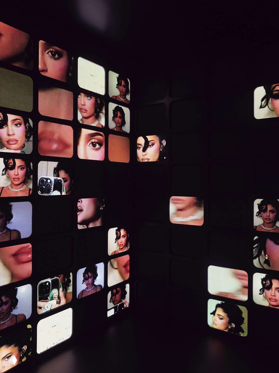 Инсталляция из множества маленьких экранов с фрагментами лиц и косметических крупным планом, затемнённая комната на вечеринке бренда.