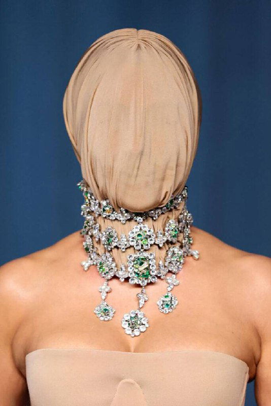 Фигура на красной дорожке с полностью закрытым бежевым чулком на голове и крупным драгоценным ожерельем с зелёными вставками.