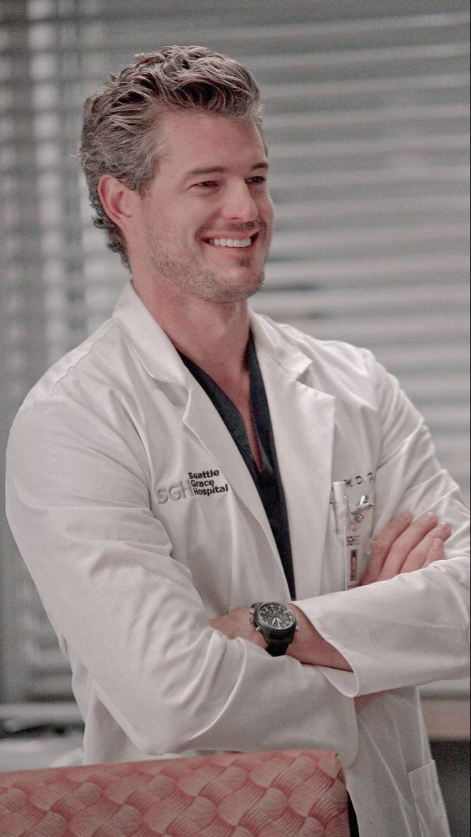 Мужчина в белом медицинском халате улыбается в интерьере больничного кабинета, видна униформа и рабочая обстановка