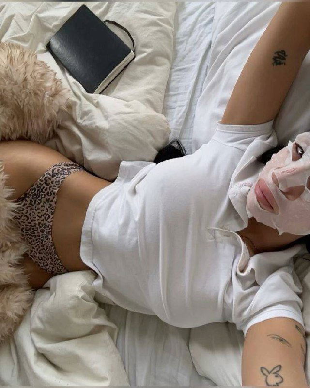 Человек лежит на кровати в тканевой маске, в белой футболке и леопардовых трусах; видна часть корпуса и тату на руке, уютная домашняя сцена.
