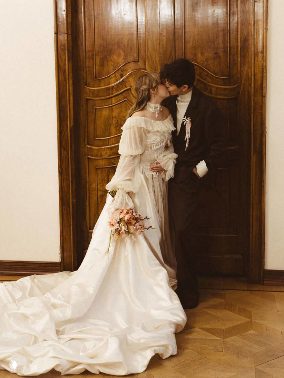 Пара целуется у резной деревянной двери: невеста в кружевном платье и букет в руках, жених в тёмном костюме, интимный кадр церемонии.