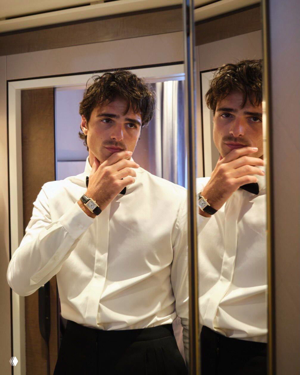 Мужчина в белой рубашке смотрит в зеркало в интерьере номера; видны двойное отражение и задумчивая поза в спокойной гамме.