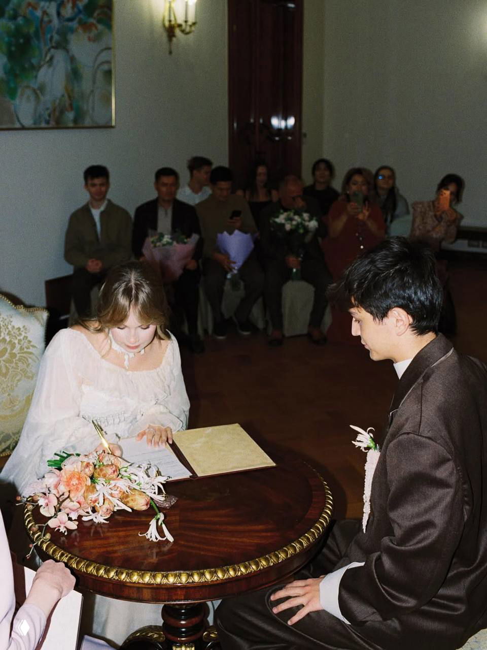 Момент подписания регистрационной книги: невеста ставит подпись за круглым резным столом, букет на столе, жених внимательно смотрит рядом.
