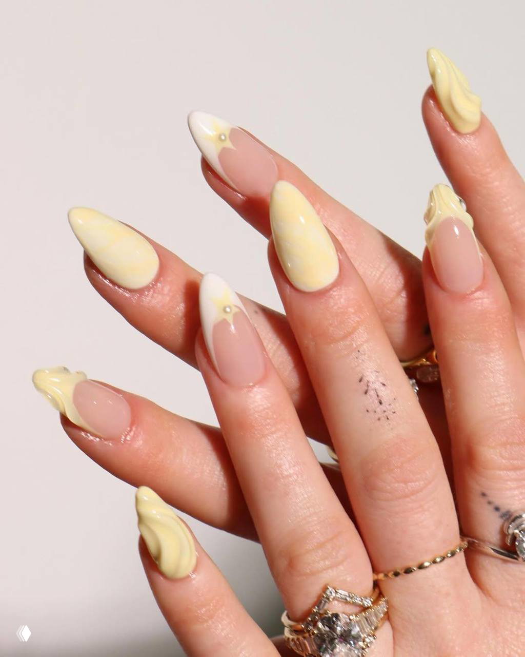 Миндалевидные ногти с кремово-жёлтым пастельным покрытием и объёмными 3D-элементами; пальцы украшены кольцами, тёплый свет.