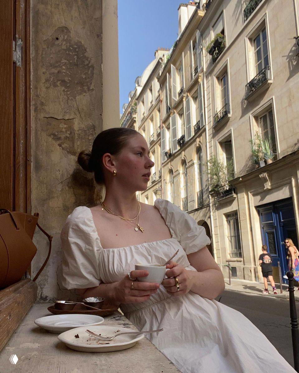 Девушка в светлом платье у окна европейской улочки с чашкой кофе в руках — мягкий уличный портрет в эстетике poetcore и vintage.