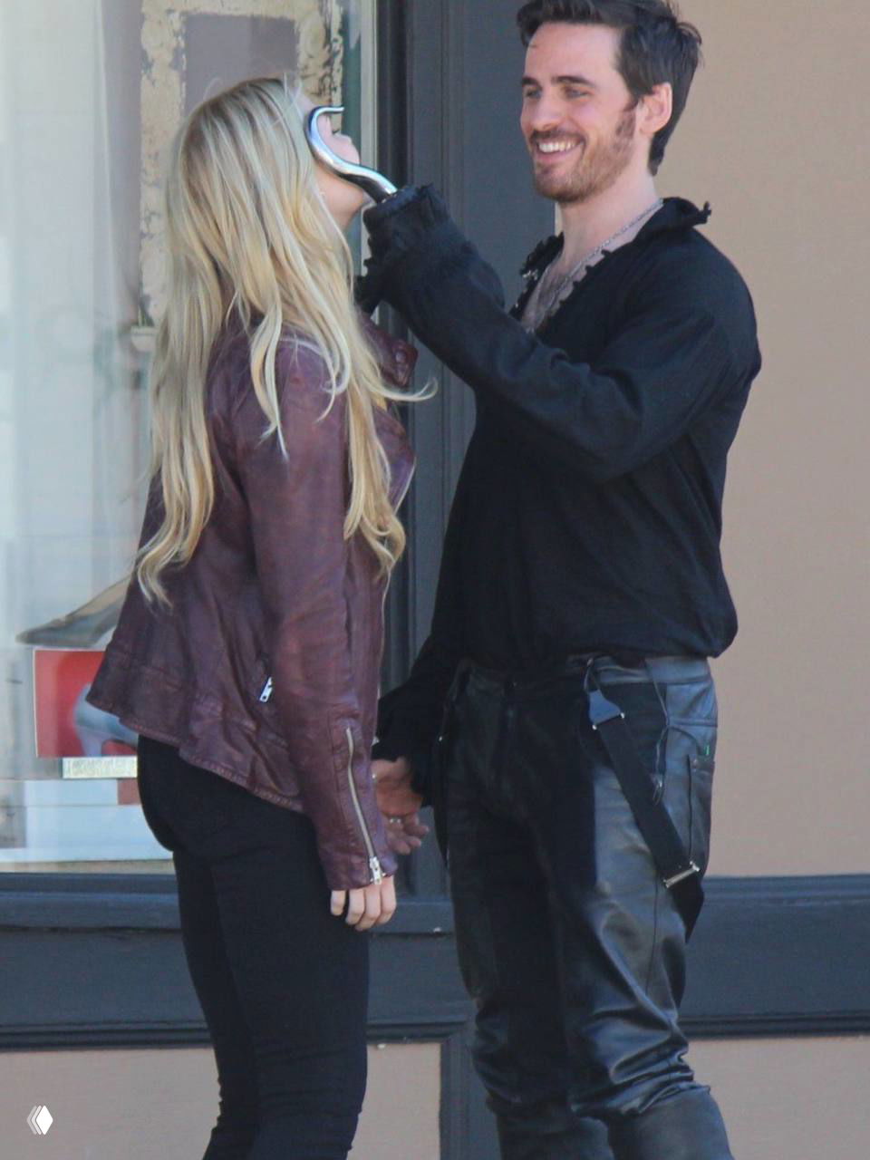 Уличная сцена со съёмок: мужчина в тёмной рубахе держит руку у лица женщины в кожаной куртке, лёгкая игровая поза на светлом фоне.