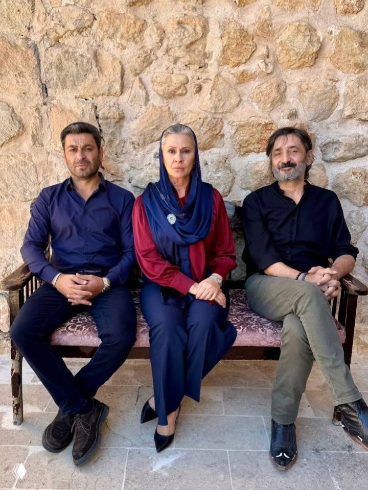 Три актёра сидят на скамейке у каменной стены в сценическом костюме: центральная женщина в шарфе и два мужчины по бокам — групповая постановочная фотография.