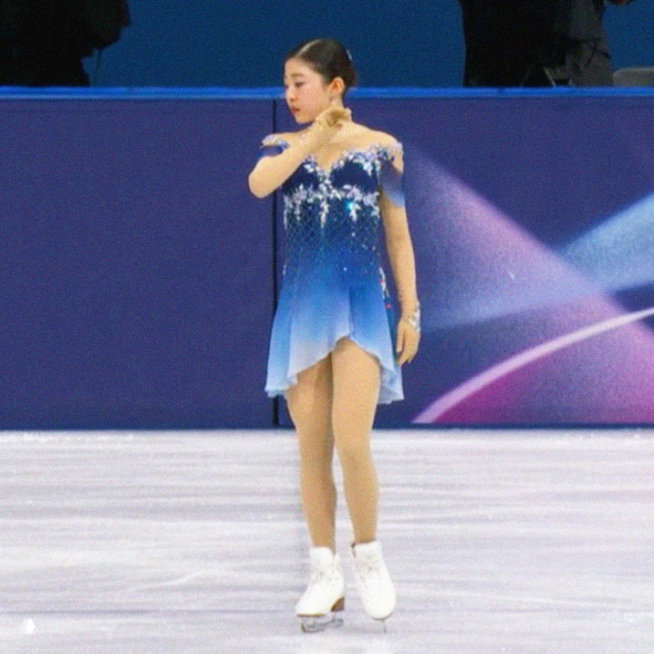 Ами Накаи в синем градиентном платье на льду во время дебютного выступления на Играх; фигуративная поза и акценты сцены видны на фоне борта катка.