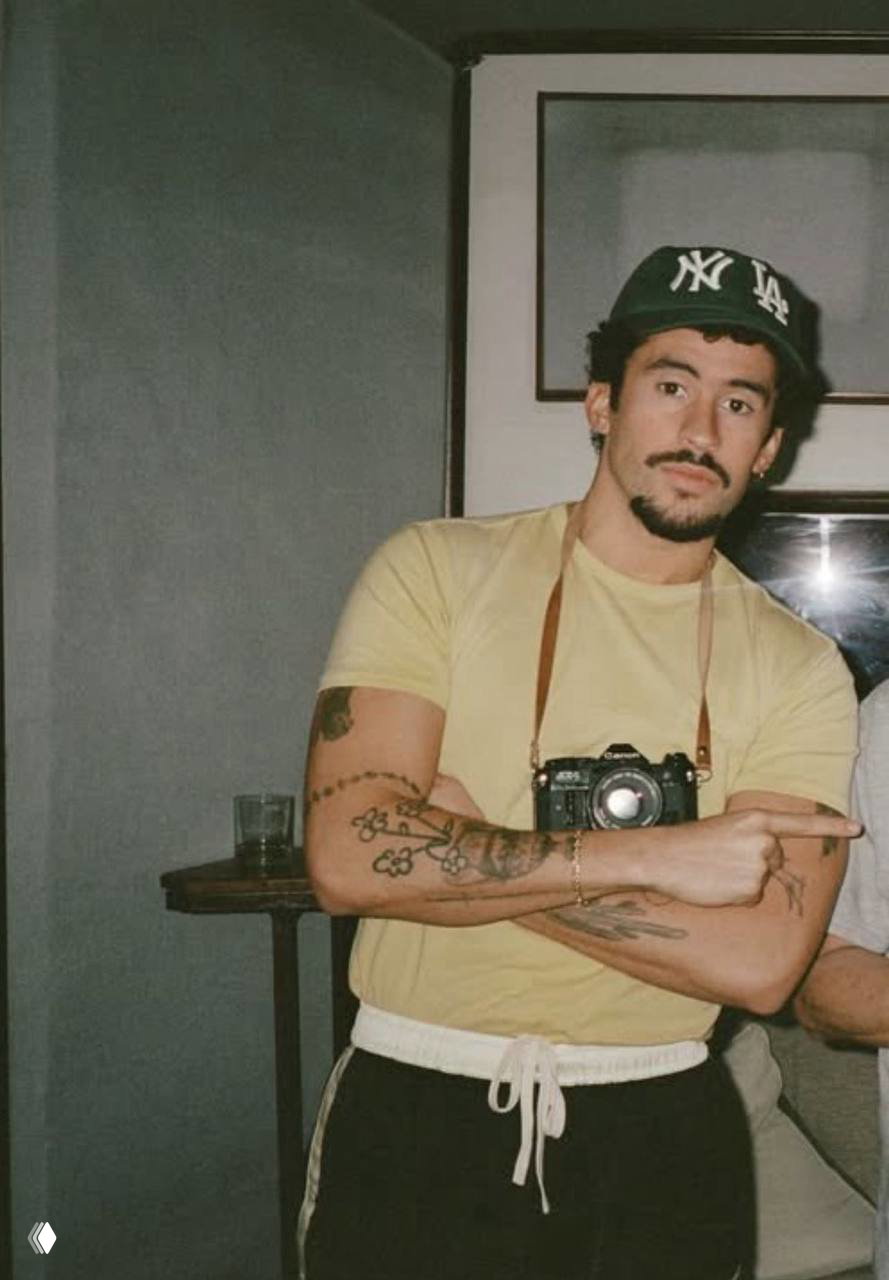 Бэд Банни на неформальной фотографии в кепке с камерой на груди, видны татуировки на руках — образ исполнителя из топ‑рейтинга Spotify.