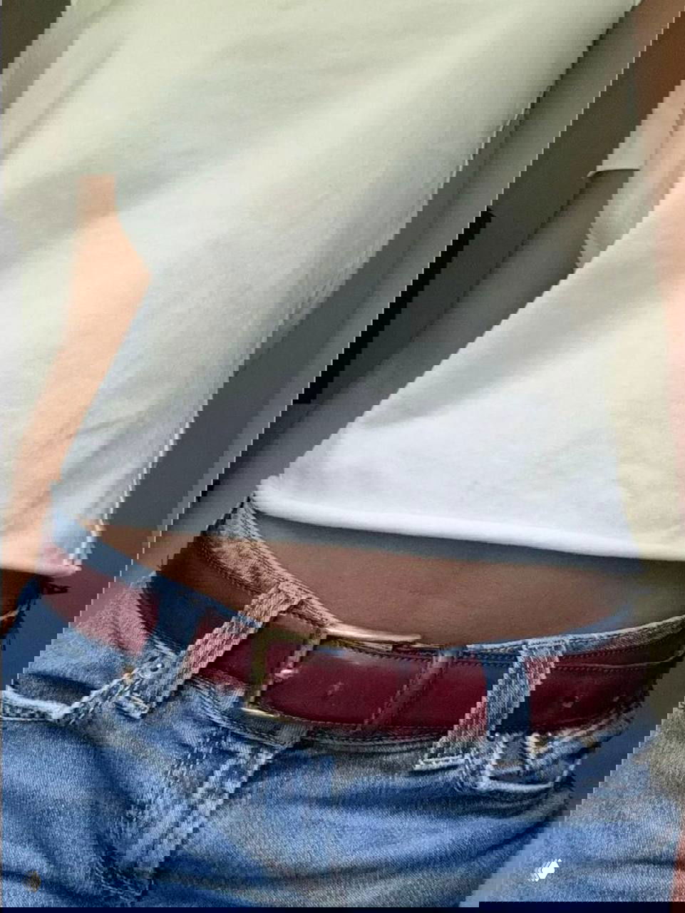 Фрагмент талии в белой футболке и тёмно-красном кожаном ремне с обычной латунной пряжкой на джинсах, нейтральный интерьер.