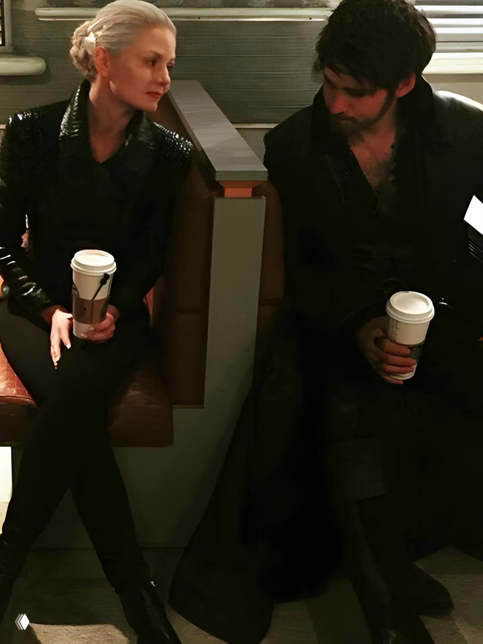 Женщина в светлом плаще и мужчина в тёмном наряде сидят рядом и улыбаются друг другу в интерьерном кадре со съёмок.