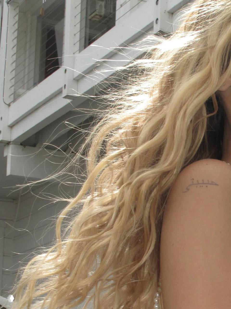 Крупный фрагмент светлых волнистых волос Беллы и часть плеча с небольшим тату, с ярким солнечным контрастом и текстурой волос.