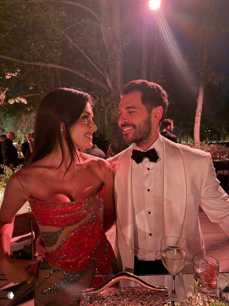 Пара на вечернем приёме: женщина в ярком платье и мужчина в смокинге улыбаются друг другу за столом на светском событии.