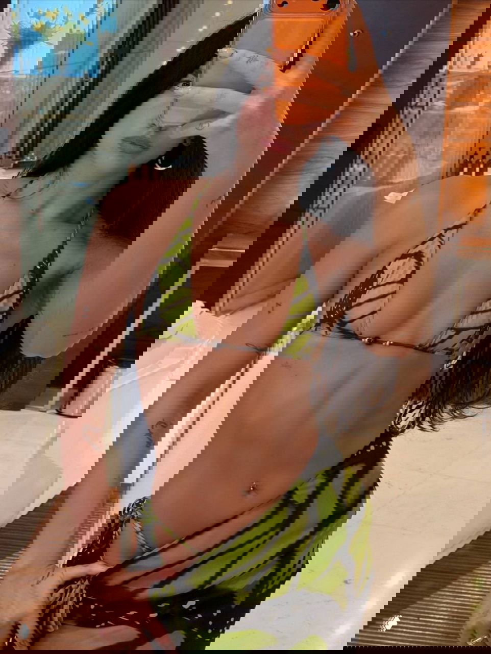 Селфи в зеркале: женщина в зелёном бикини с принтом крыла бабочки на фоне номера отеля; видны детали ванной и интерьер.