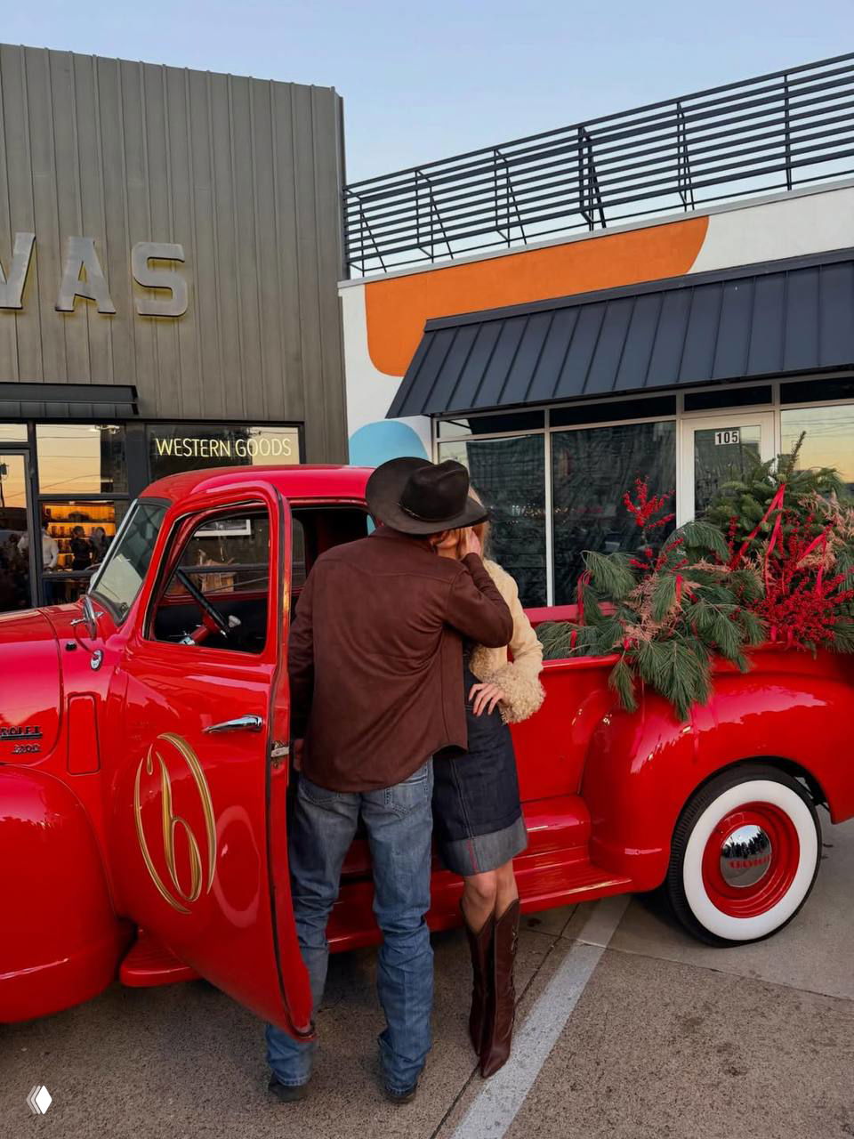 Пара обнимается у открытой двери красного пикапа перед витриной магазина; вечерняя уличная обстановка.