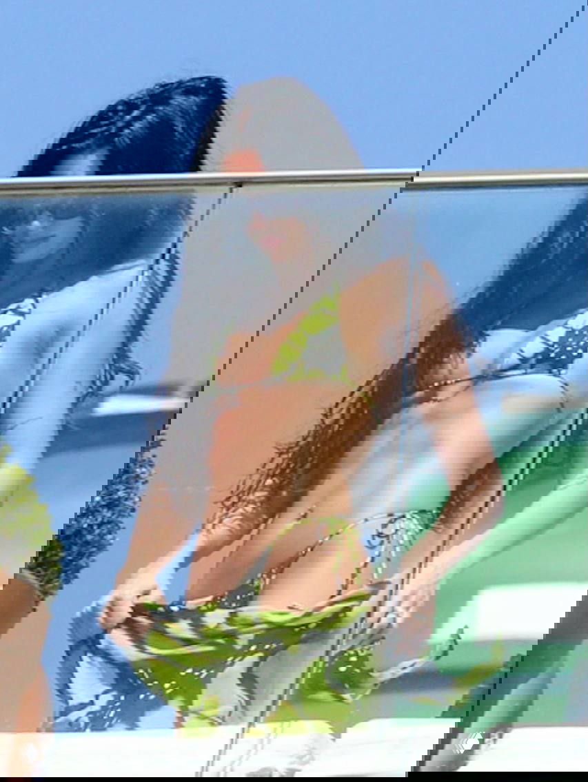 Женщина в зелёном бикини с принтом аккуратно поправляет парео на террасе; на заднем плане небо и морской пейзаж.