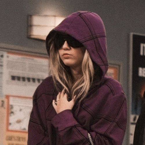 Женщина в тёмно‑фиолетовом худи и тёмных очках на фоне доски объявлений; винтажная обработка кадра, интерьер сцены ситкома.