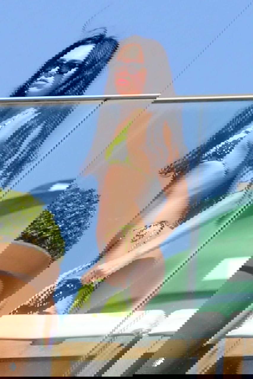 Фигура в зелёном бикини у стеклянного ограждения на пляжной террасе; солнечный свет подчёркивает контуры и силуэт.