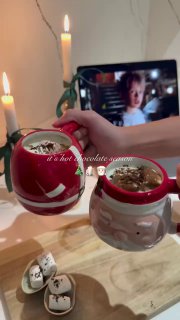 Две праздничные кружки с горячим шоколадом и маршмеллоу на деревянном столе; в фоне зажжённые свечи и экран с фильмом.