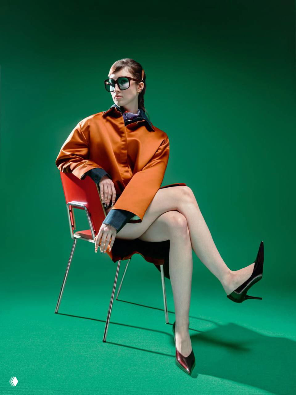 Портрет Лизы Барашек в оранжевом плаще на зелёном фоне: сидящая поза на стуле, очки и журнальная постановка съёмки для интервью.