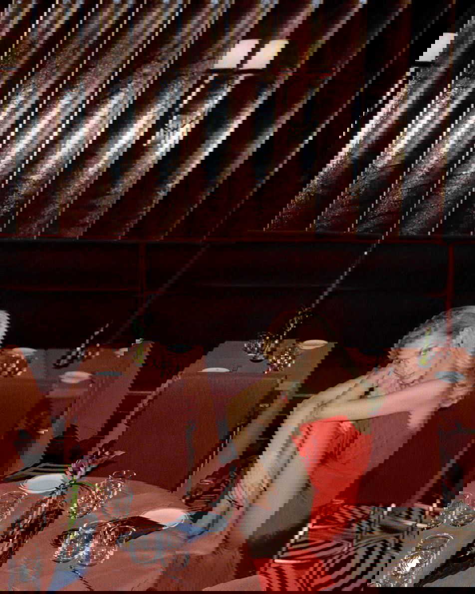 Модель в красном платье сидит в ресторане в стиле 80‑х: тёплые тона, винтажная мебель и атмосферная интерьерная съёмка в глянцевом ключе.