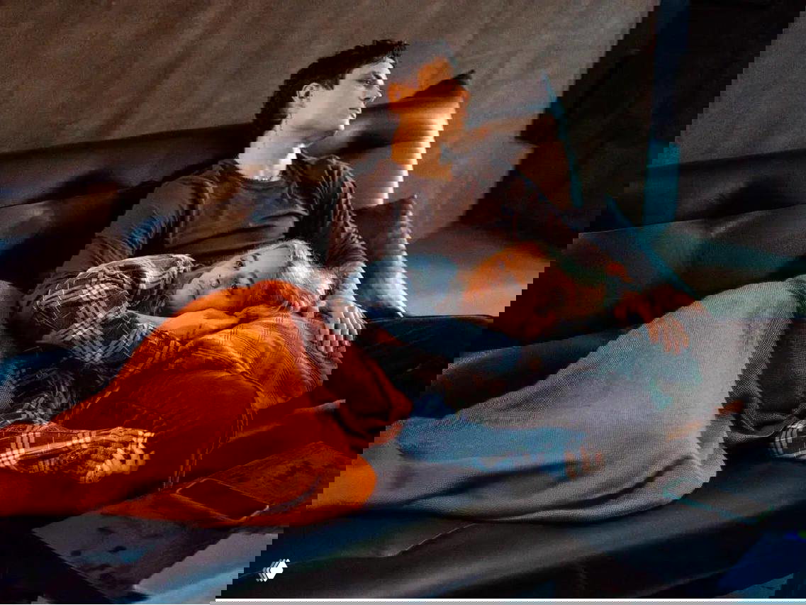 Интимный кадр: парень и девушка лежат на диване под оранжевым пледом, актёры обнимаются в домашней, уютной обстановке.