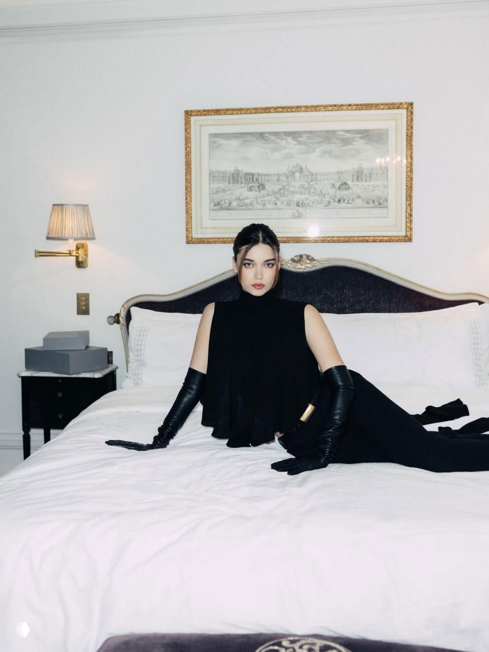 Афра Сарачоглу на кровати в total black от Balenciaga: свободный топ с открытой спиной, длинные чёрные перчатки и интерьерная съёмка в парижском отеле.