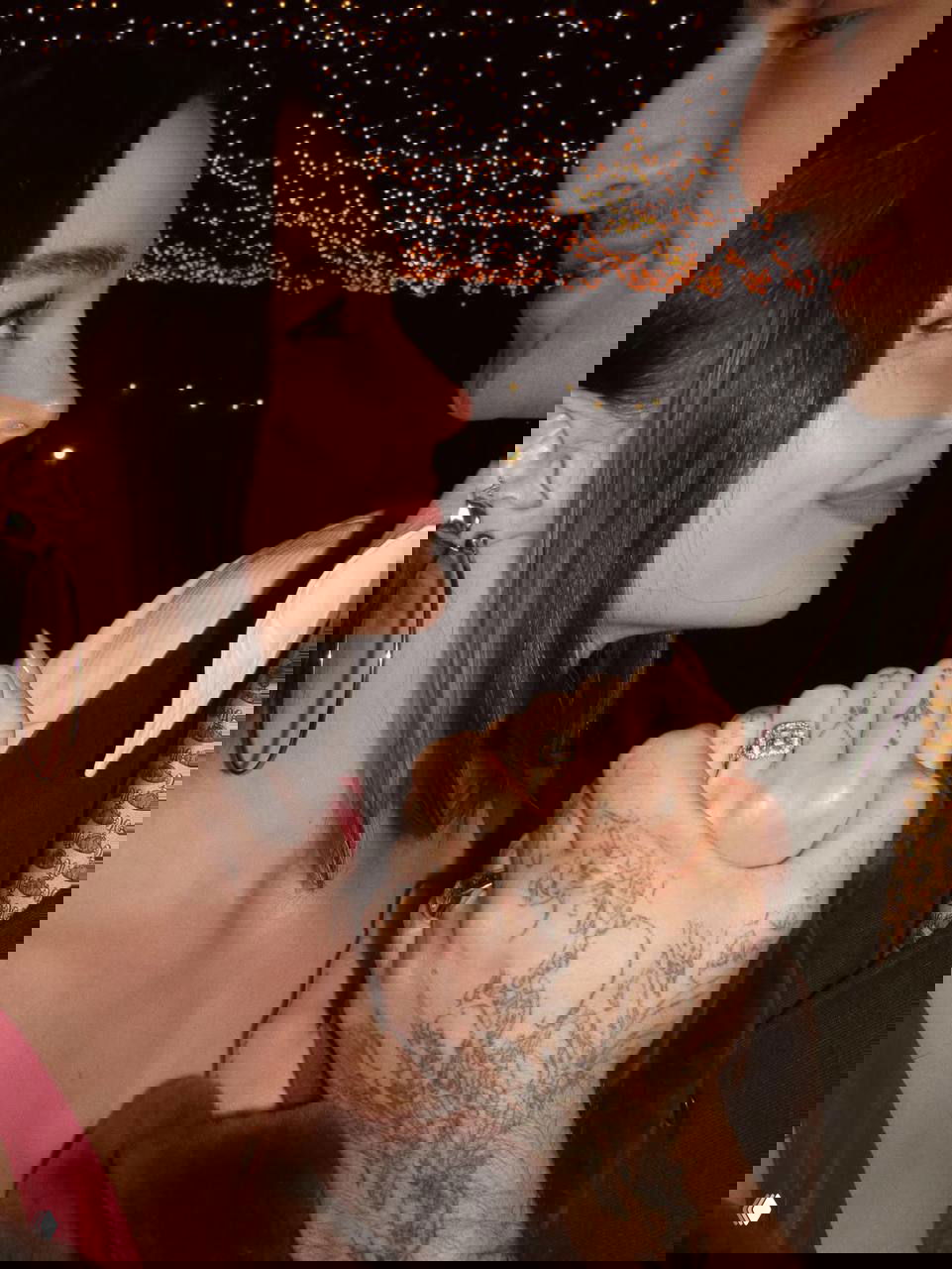 Крупный план рук с кольцом и татуировками: Дав Кэмерон и Дамиано улыбаются на фоне огней, видно обручальное кольцо.