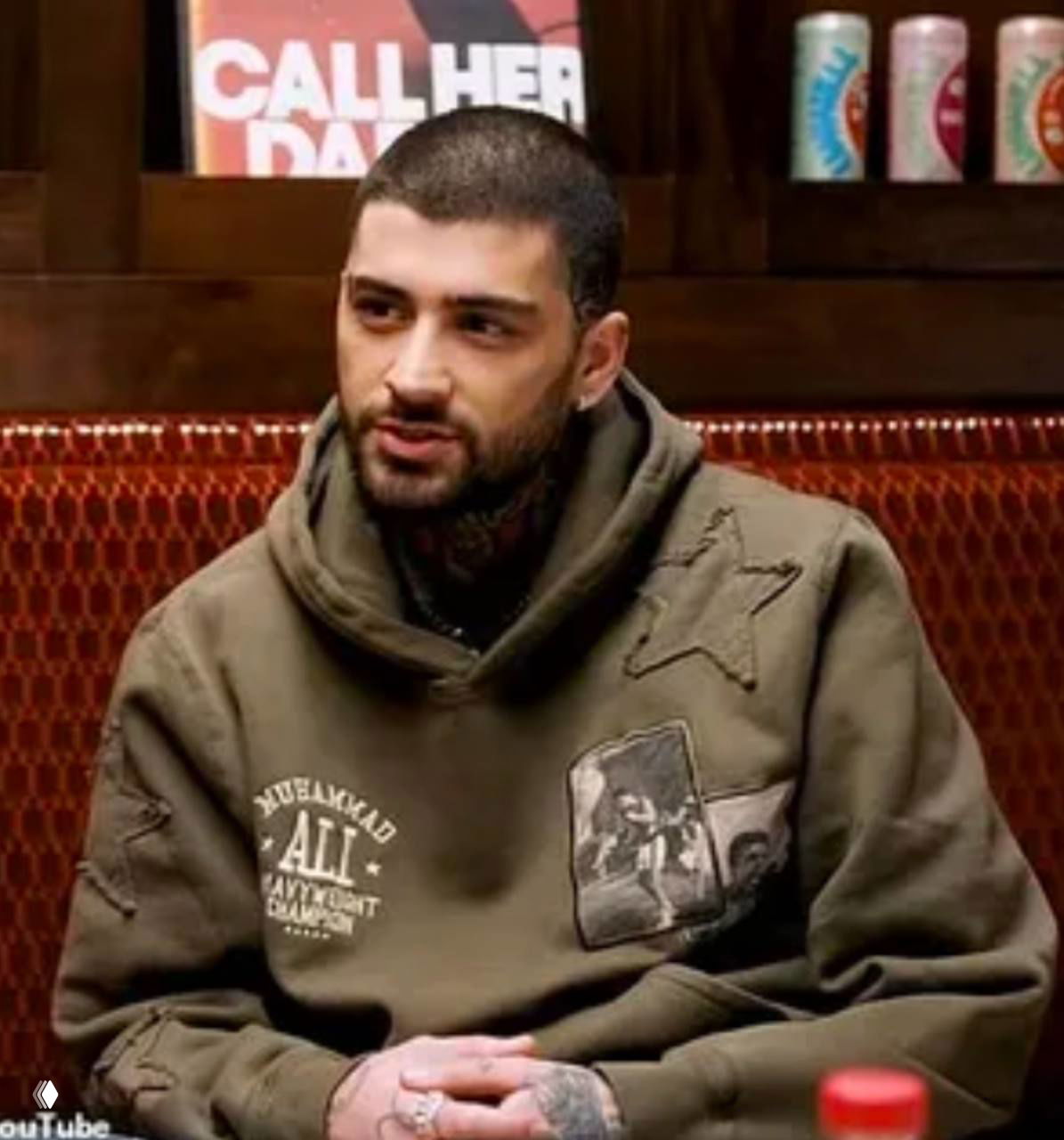 Зейн Малик в худи во время интервью в студии Call Her Daddy: артист сидит за столом, на заднем плане видна вывеска шоу и напитки на полках.