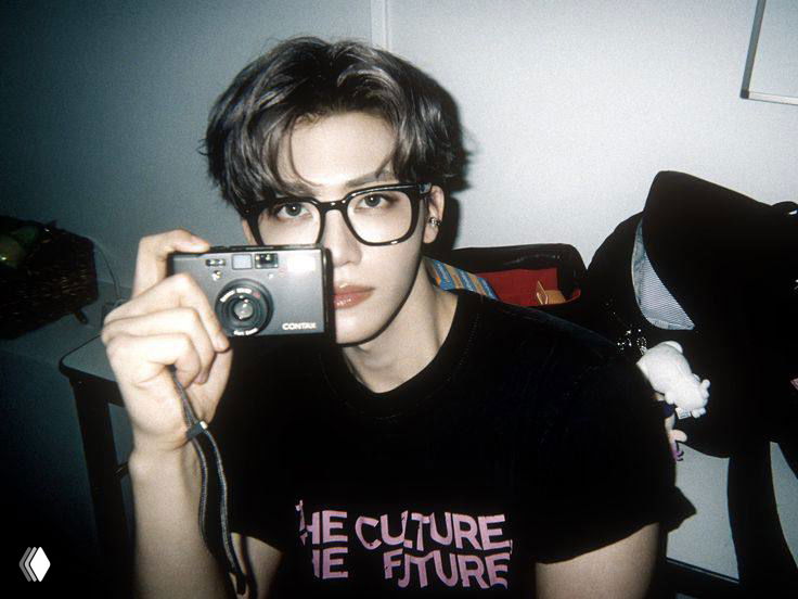 Айдол в очках держит ручную пленочную камеру, сидя в комнате; фото с мягким контрастом и ретро-атмосферой.