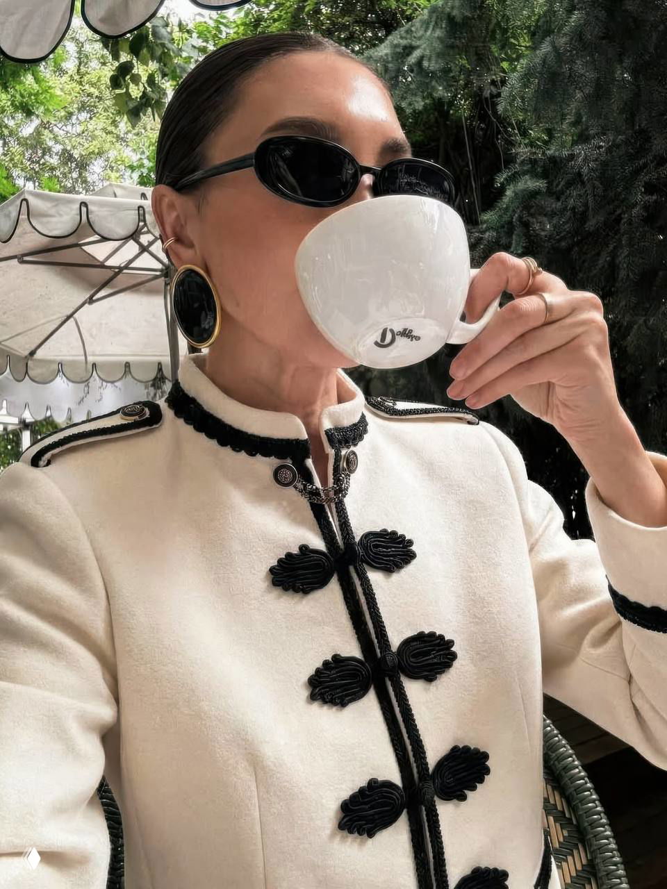 Крупный план белого гусарского жакета с чёрной окантовкой: аксессуары, очки и чашка кофе в уличном кафе — акцент на отделке и деталях.