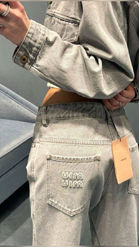 Серые джинсы крупным планом в примерочной: пояс, бирка и карман на фоне мягкого освещения, подчёркнутая фактура ткани.