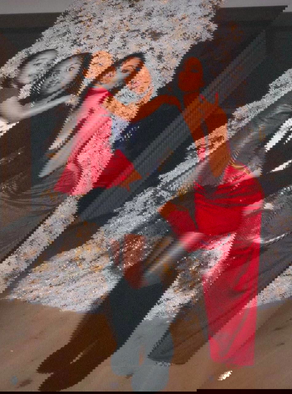 Семейная фотография у украшенной рождественской ёлки: взрослая женщина и две девочки в праздничных нарядах позируют вместе, тёплая композиция в интерьере.