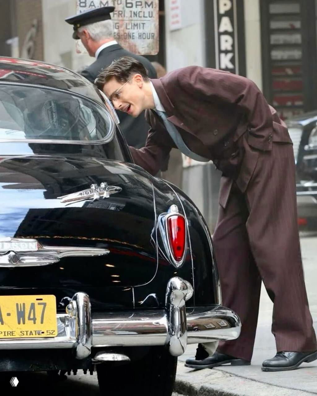 Сцена с мужчиной, наклонившимся к ретро-автомобилю на городской улице; классический костюм и атмосфера съёмочного кадра середины века.