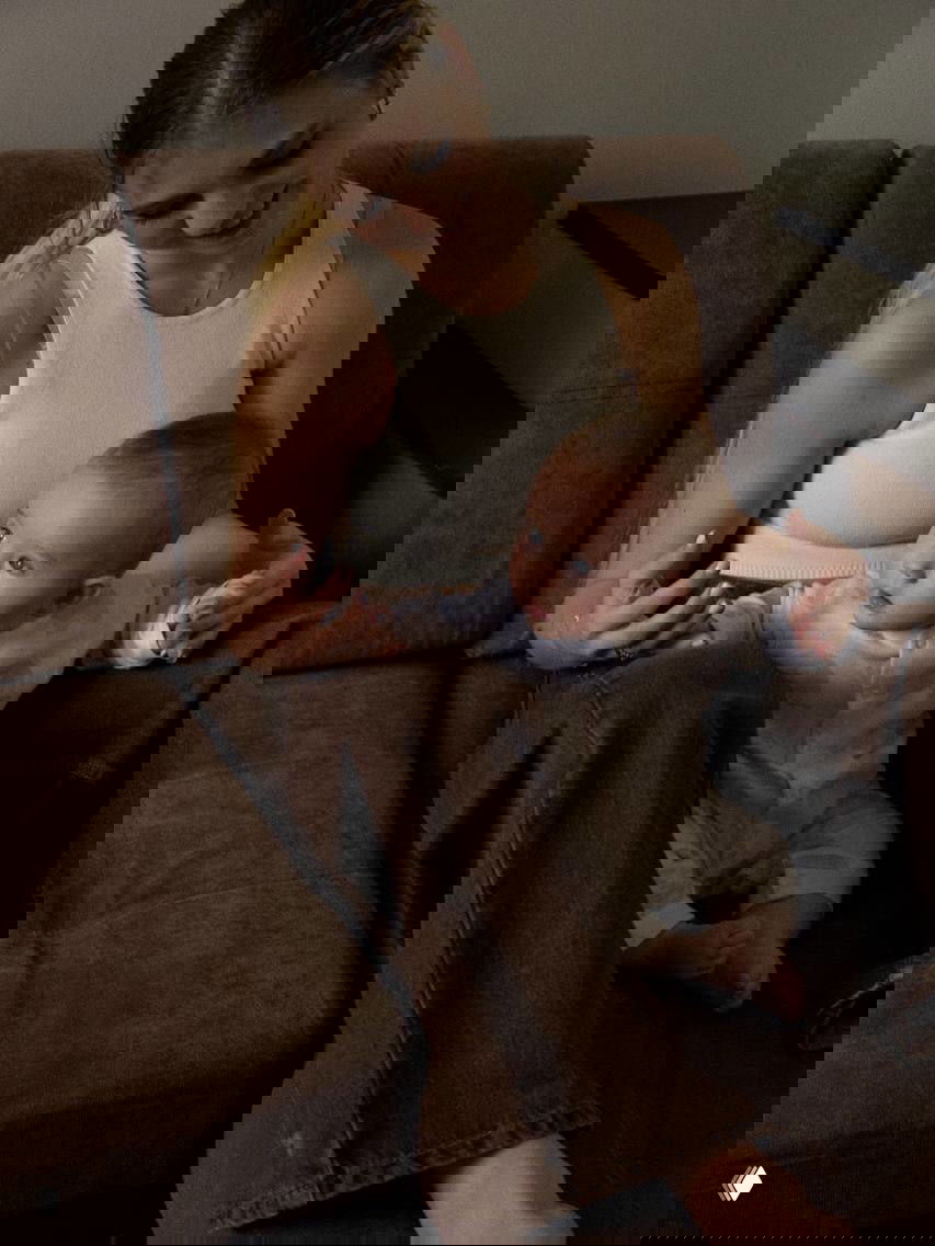 Спортсменка улыбается, поддерживая ребёнка на коленях; снимок показывает взаимодействие и первые жесты малыша в уютной комнате с коричневым диваном.