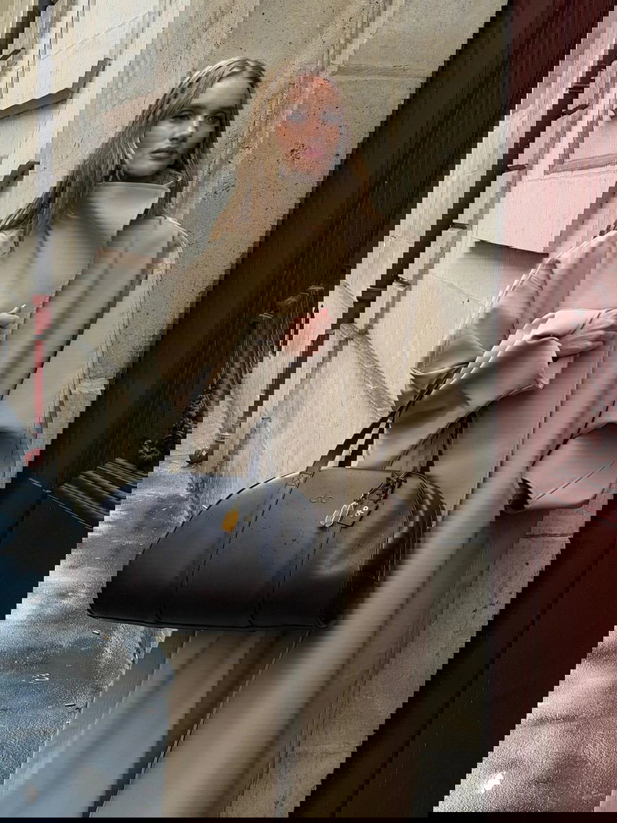 Модель в бежевом плаще с высоким воротником-стойкой держит коричневую сумку на фоне каменного фасада городской улицы, образ в спокойной палитре.