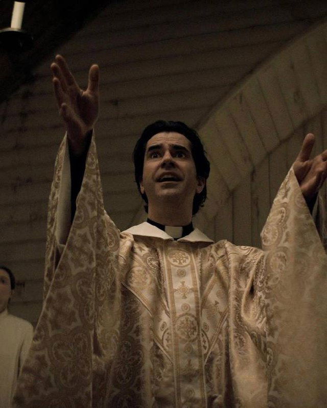 Кадр с мужчиной в церковном облачении, поднявшим руки в жесте проповеди на тёмном фоне; религиозный мотив в контексте хоррора и напряжённая сценография.