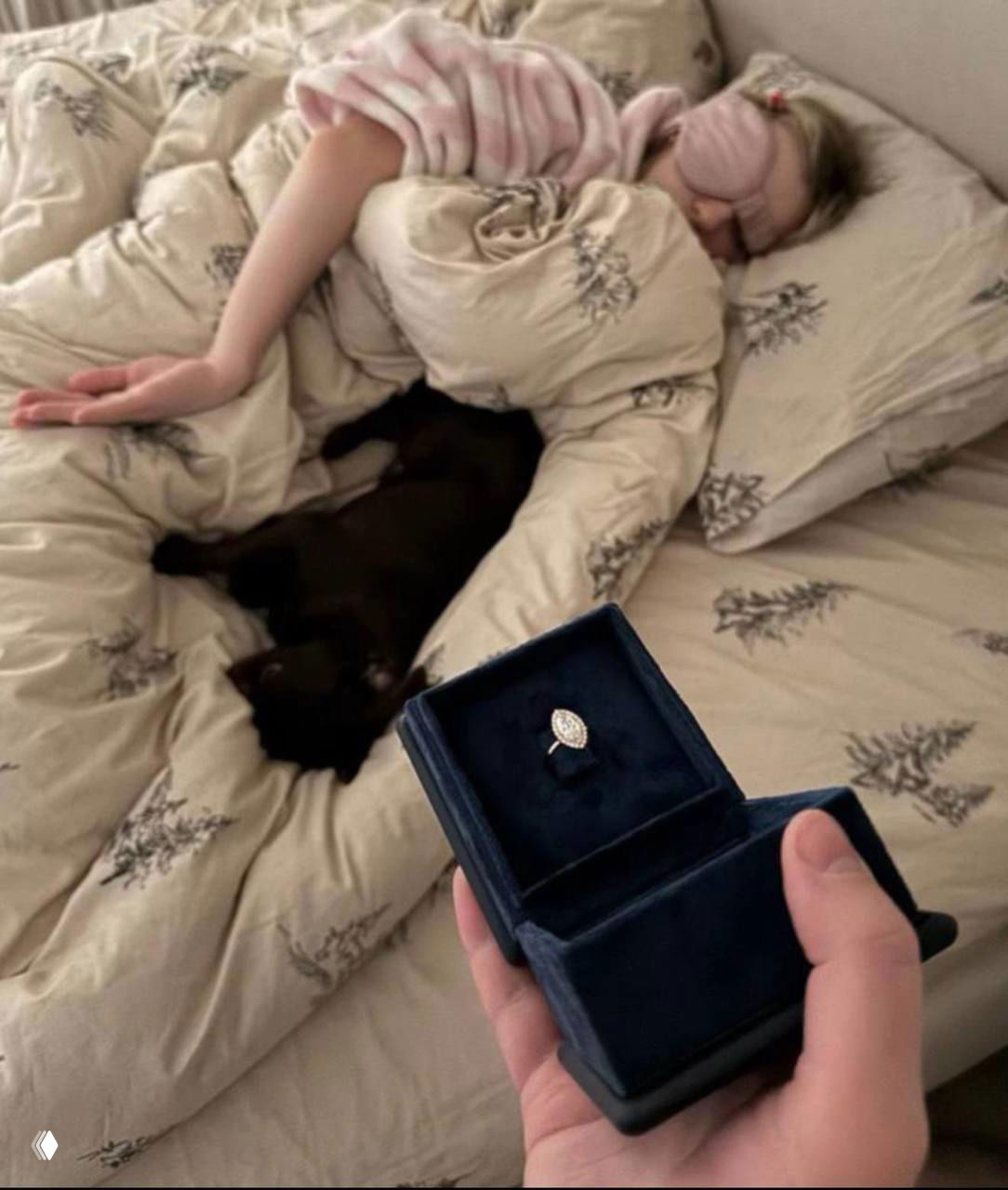 Крупный план раскрытой бархатной коробочки с кольцом на переднем плане; на заднем плане на постели спящий человек и постельное бельё.