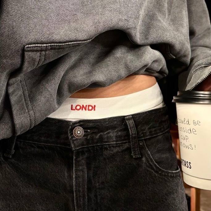 Фрагмент талии в заправленных джинсах с видимой биркой 'LONDI' и бумажным стаканом кофе в руке; небрежная повседневная эстетика улицы.