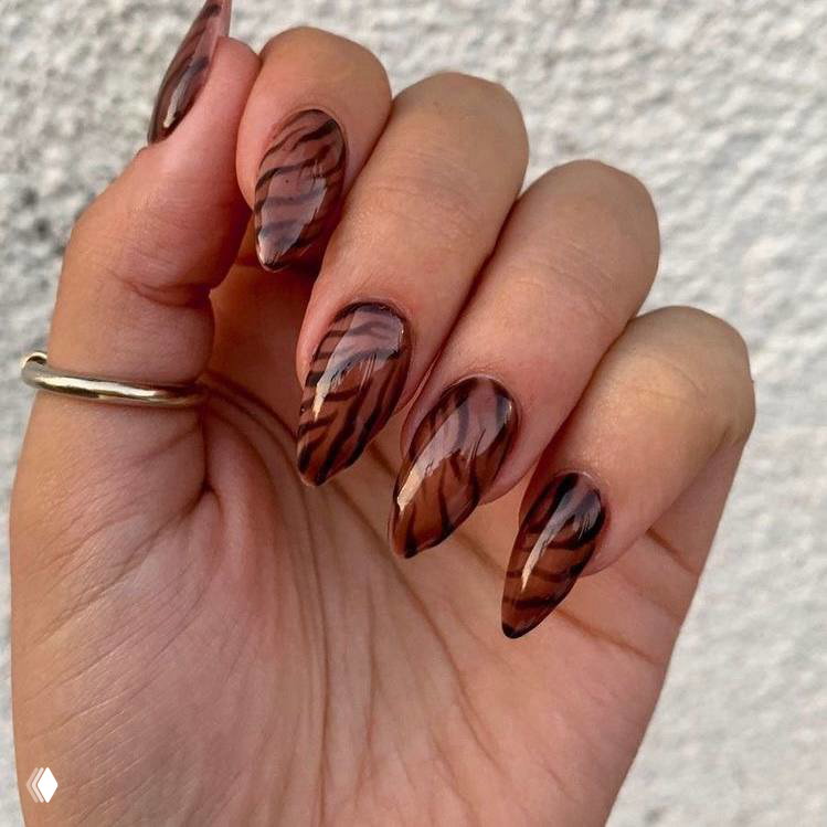 Рука с удлинёнными ногтями в тёплых коричневых тонах, выполненных волнистыми зебровыми/тигриными полосами в прозрачном и глянцевом финише.